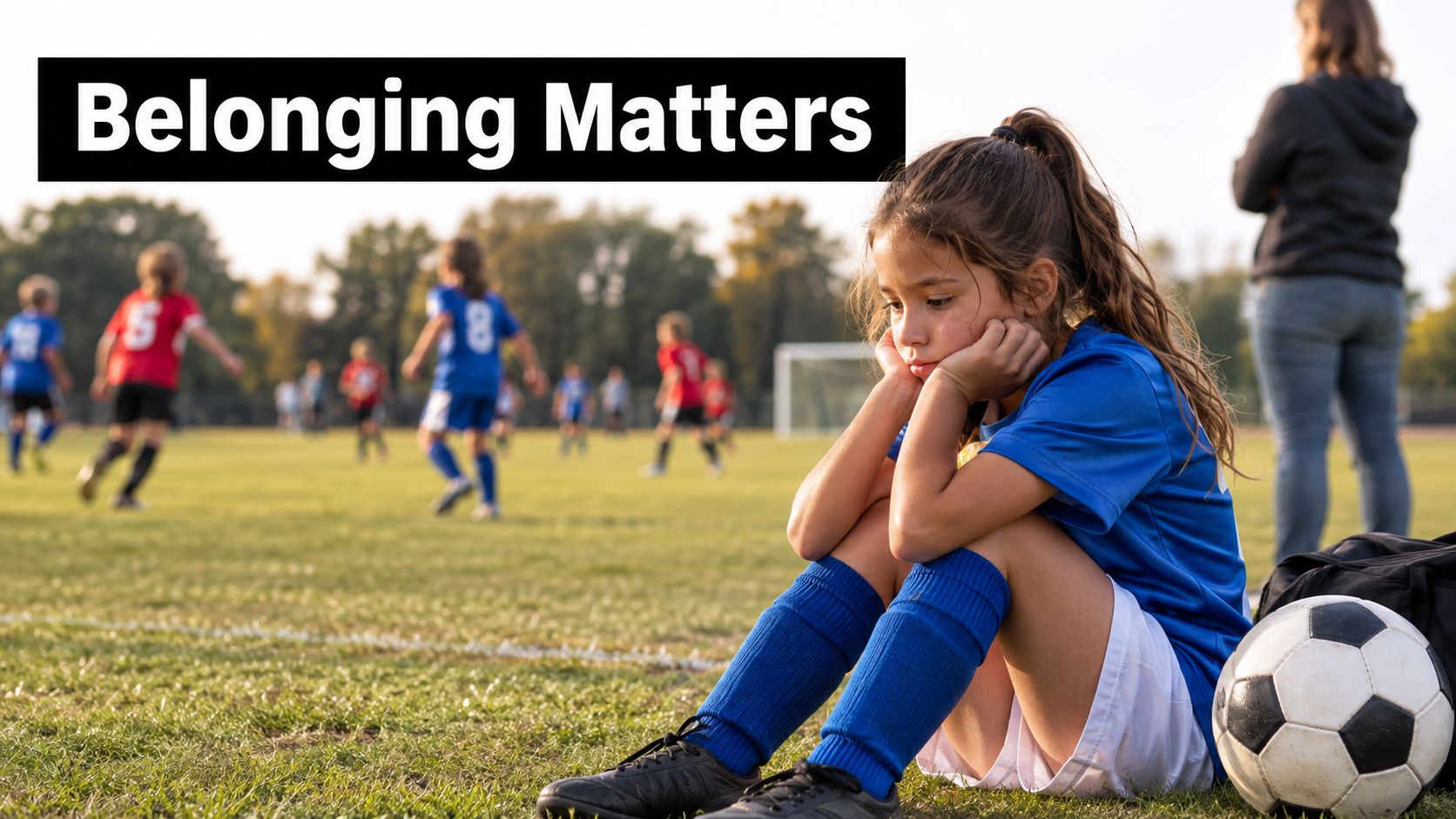 A sad young girl in a blue soccer uniform sitting alone on a field while others play.