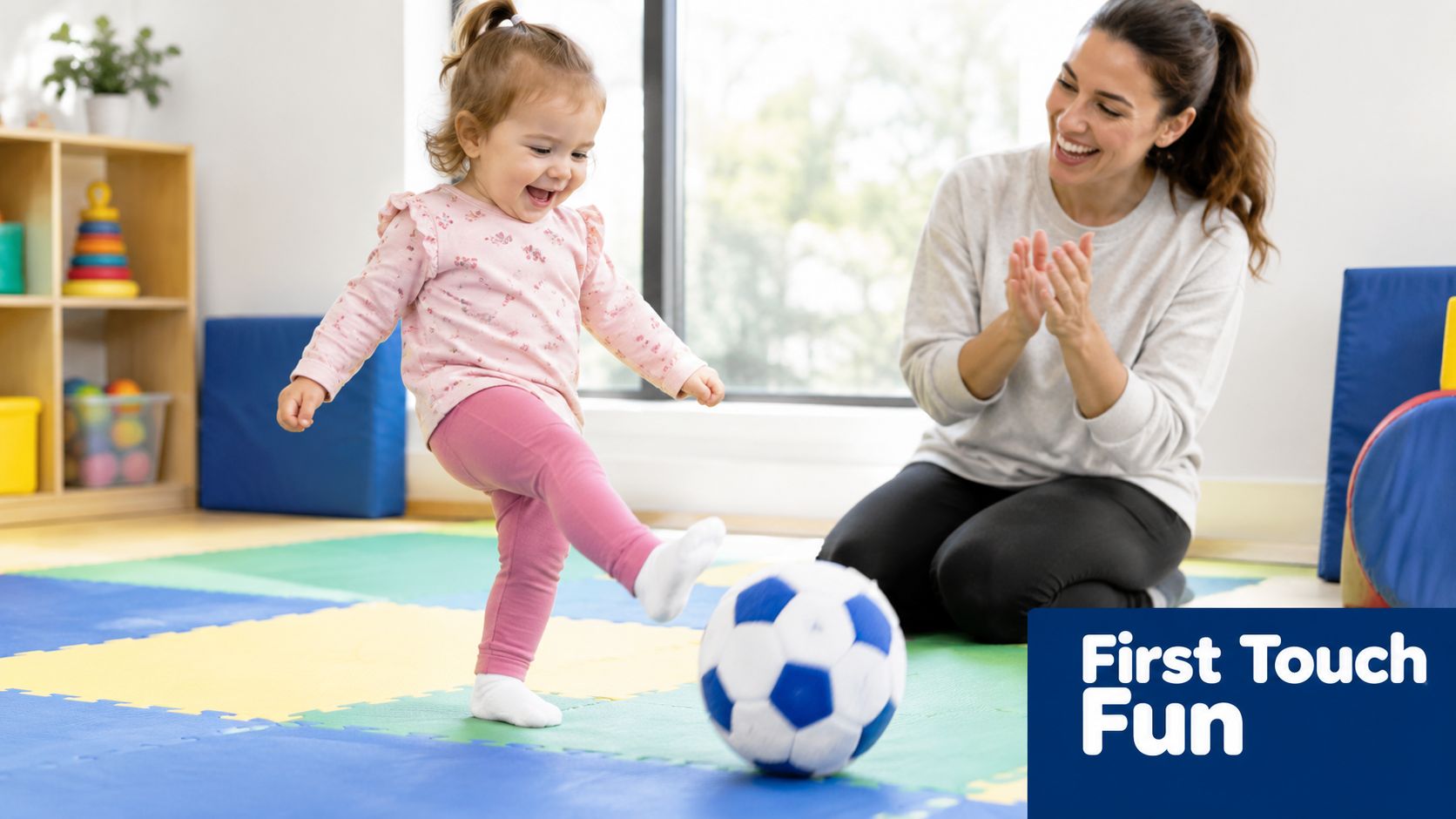A happy toddler girl kicking a soccer ball while her mother claps in a brightly lit indoor playroom.