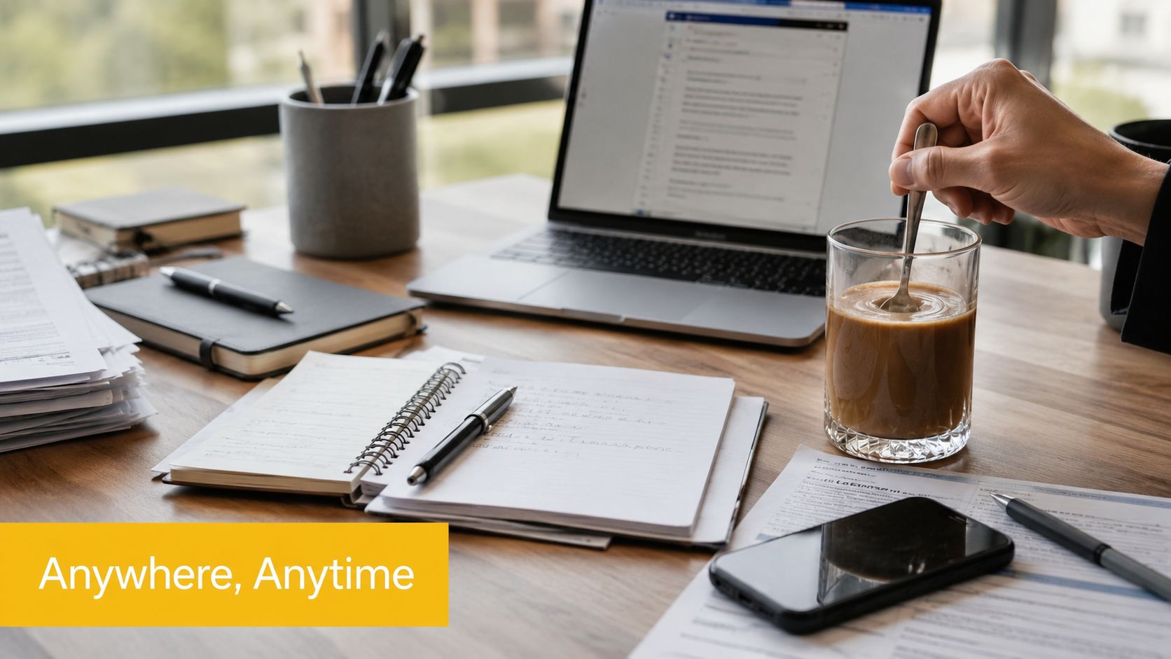A professional office desk with a laptop, notebook, pens, a smartphone, and a person stirring coffee.