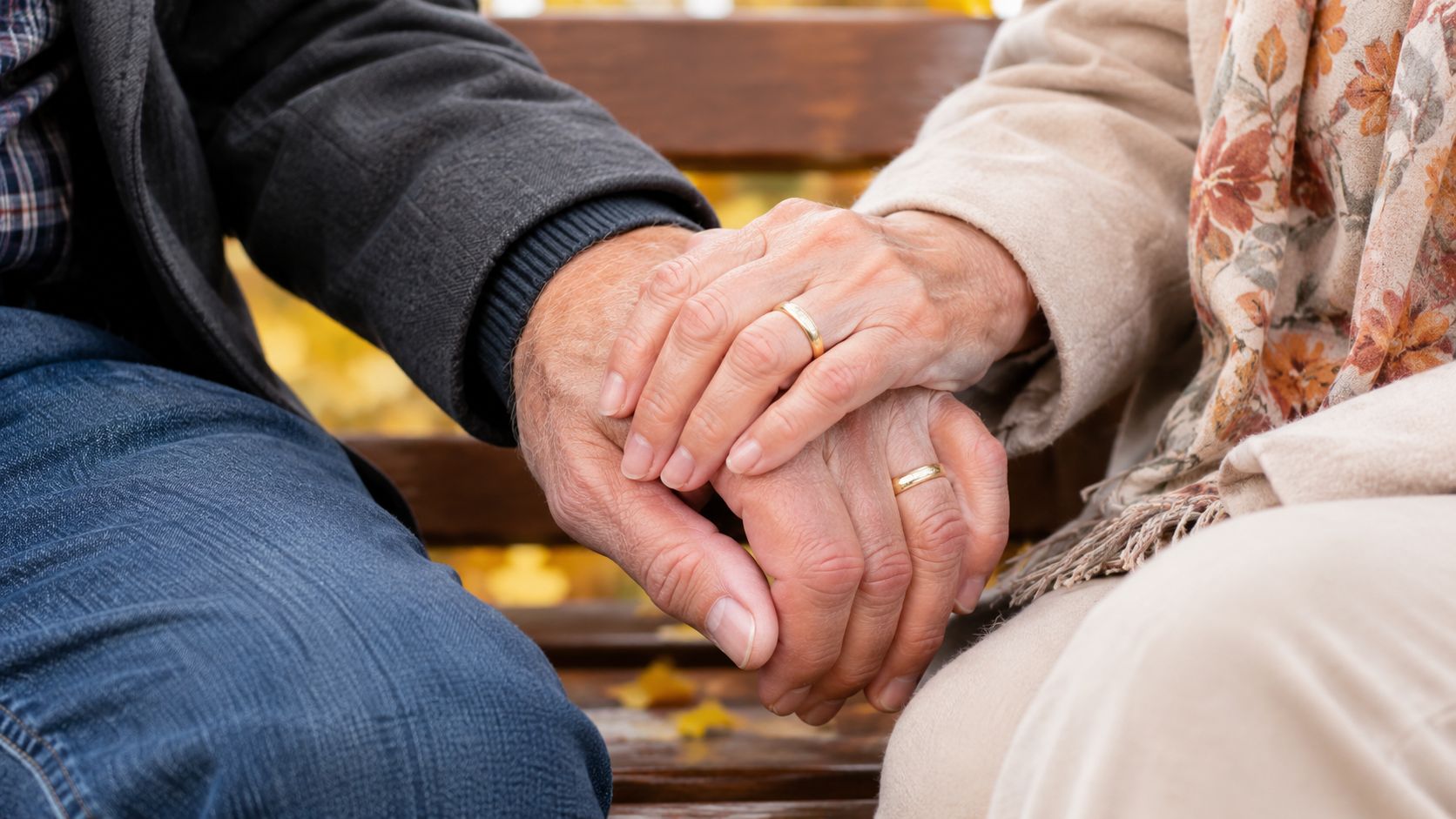 An elderly couple holding hands on a park bench during autumn, showcasing their golden wedding rings.