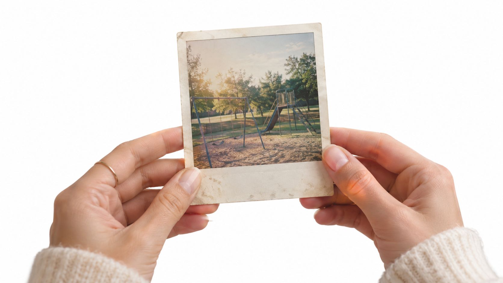 A person holding an old, vintage-style polaroid photograph showing an empty children's playground at sunset.