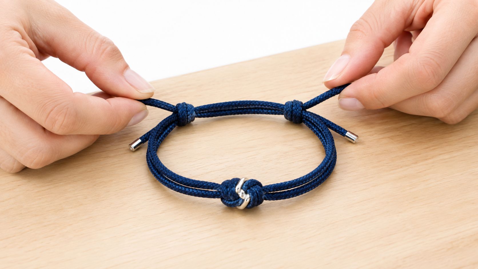 Zwei Hände halten ein blaues Segeltau-Armband mit einem silbernen Knoten und verstellbaren Enden auf einer Holzoberfläche.