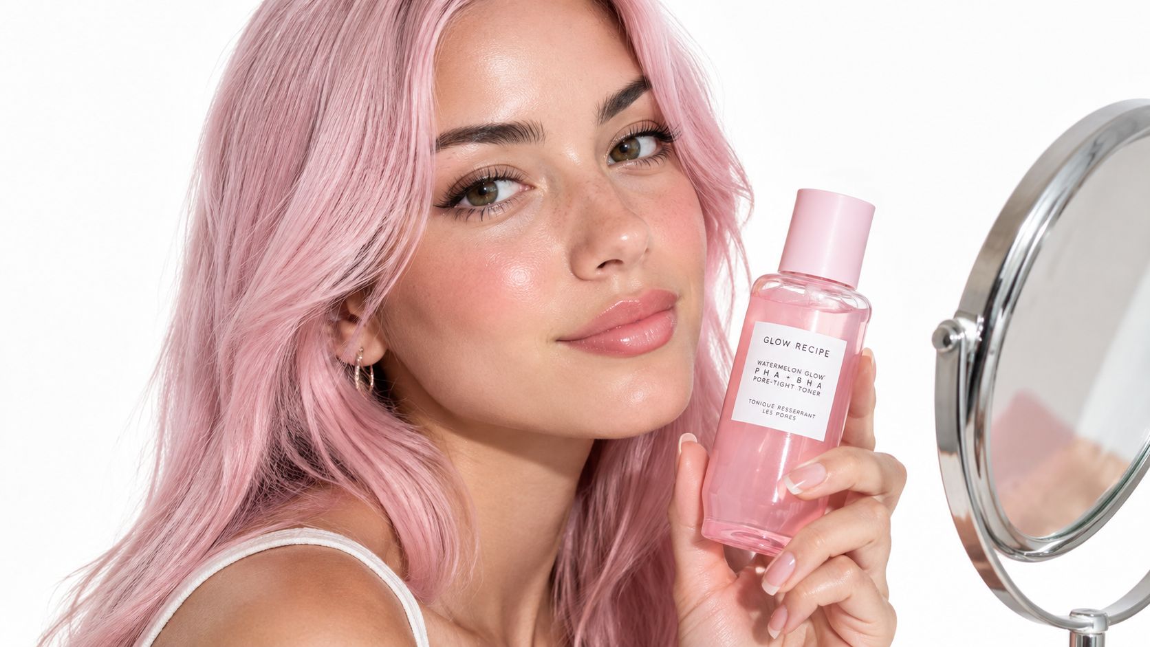 A woman with pastel pink hair holding a bottle of Glow Recipe watermelon toner by a mirror.