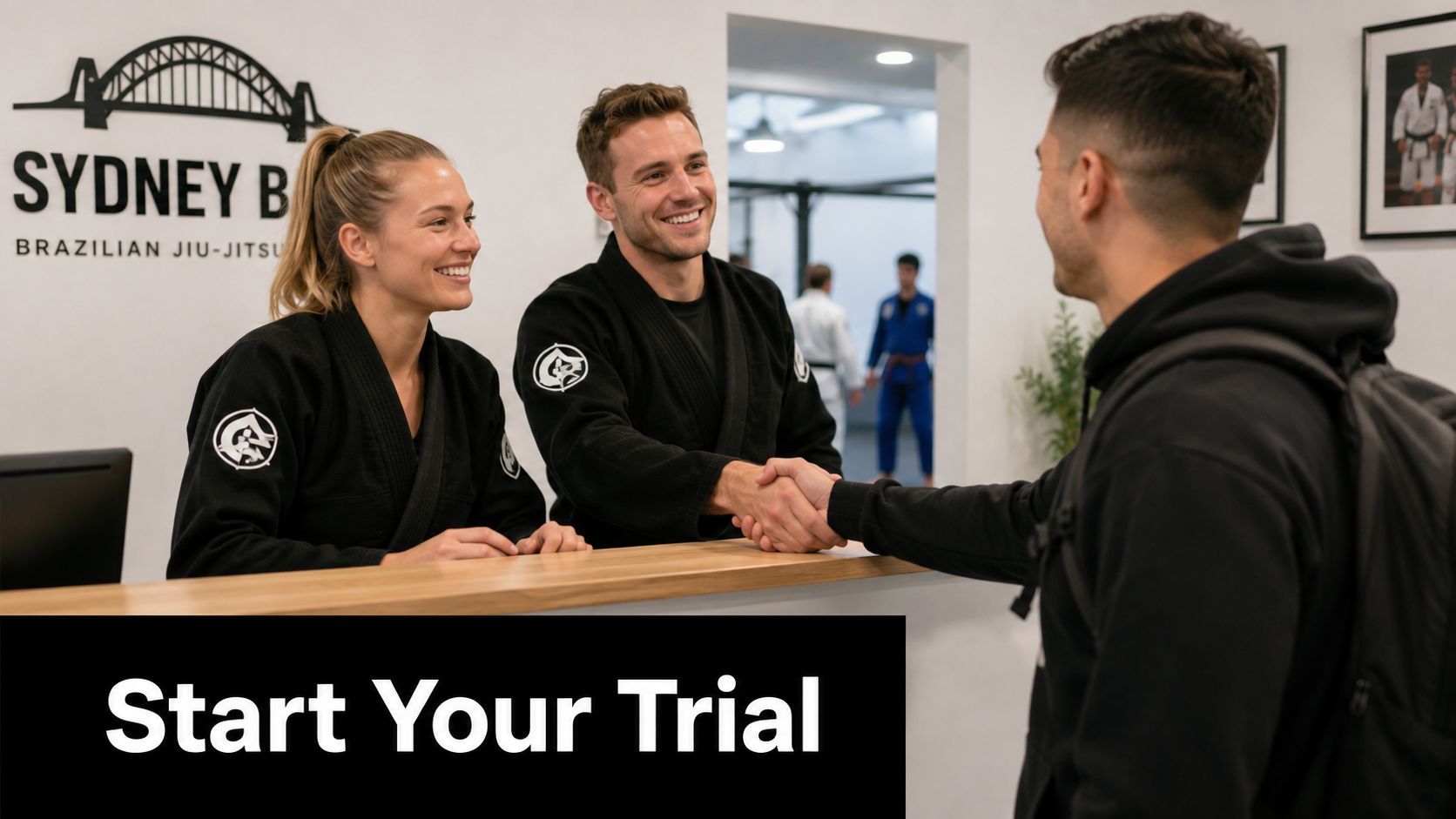 A woman and man in black jiu-jitsu uniforms greeting a new student at a gym reception desk.