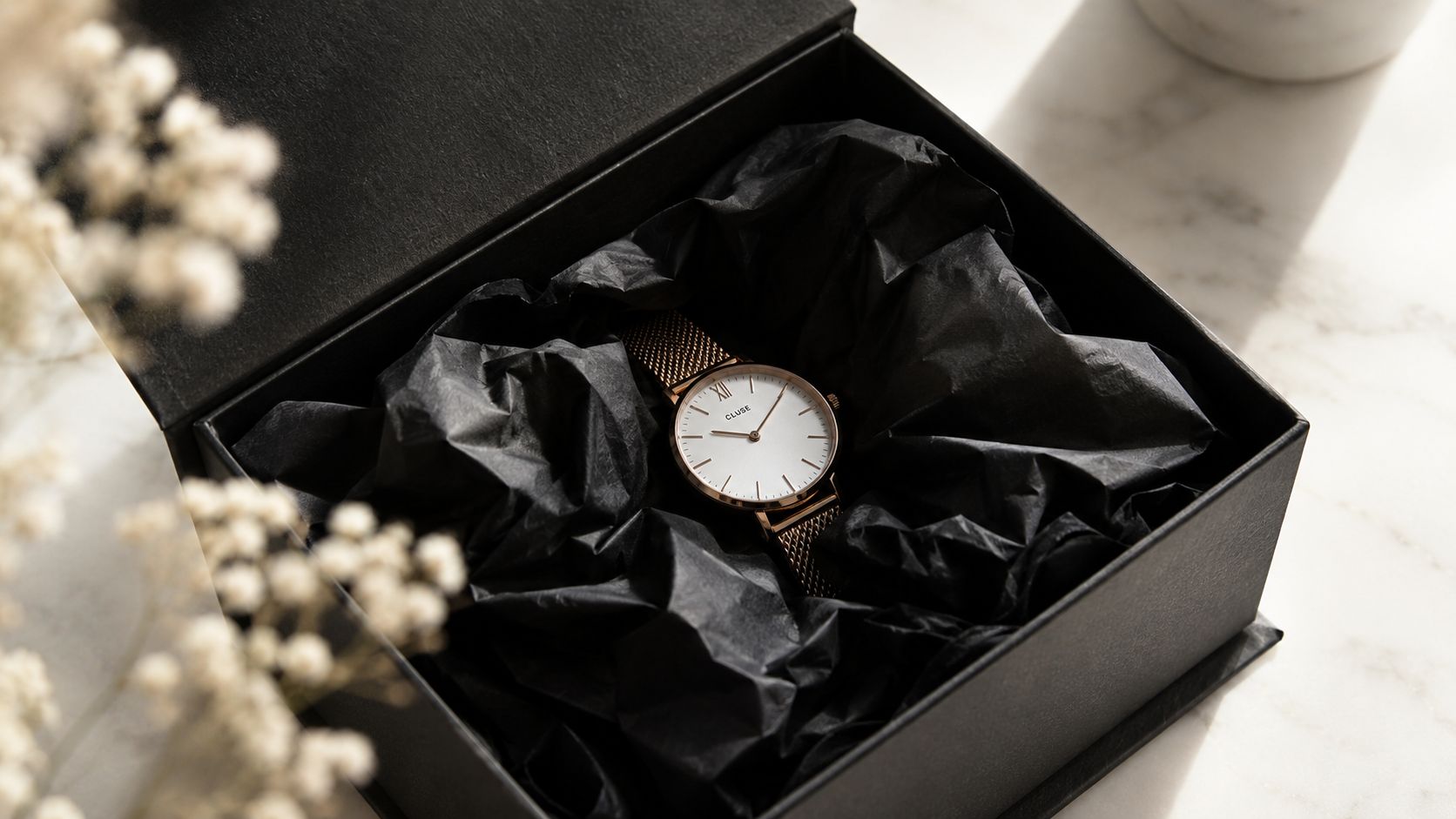 A elegant gold Cluse wristwatch nestled inside a black box lined with crumpled black tissue paper.