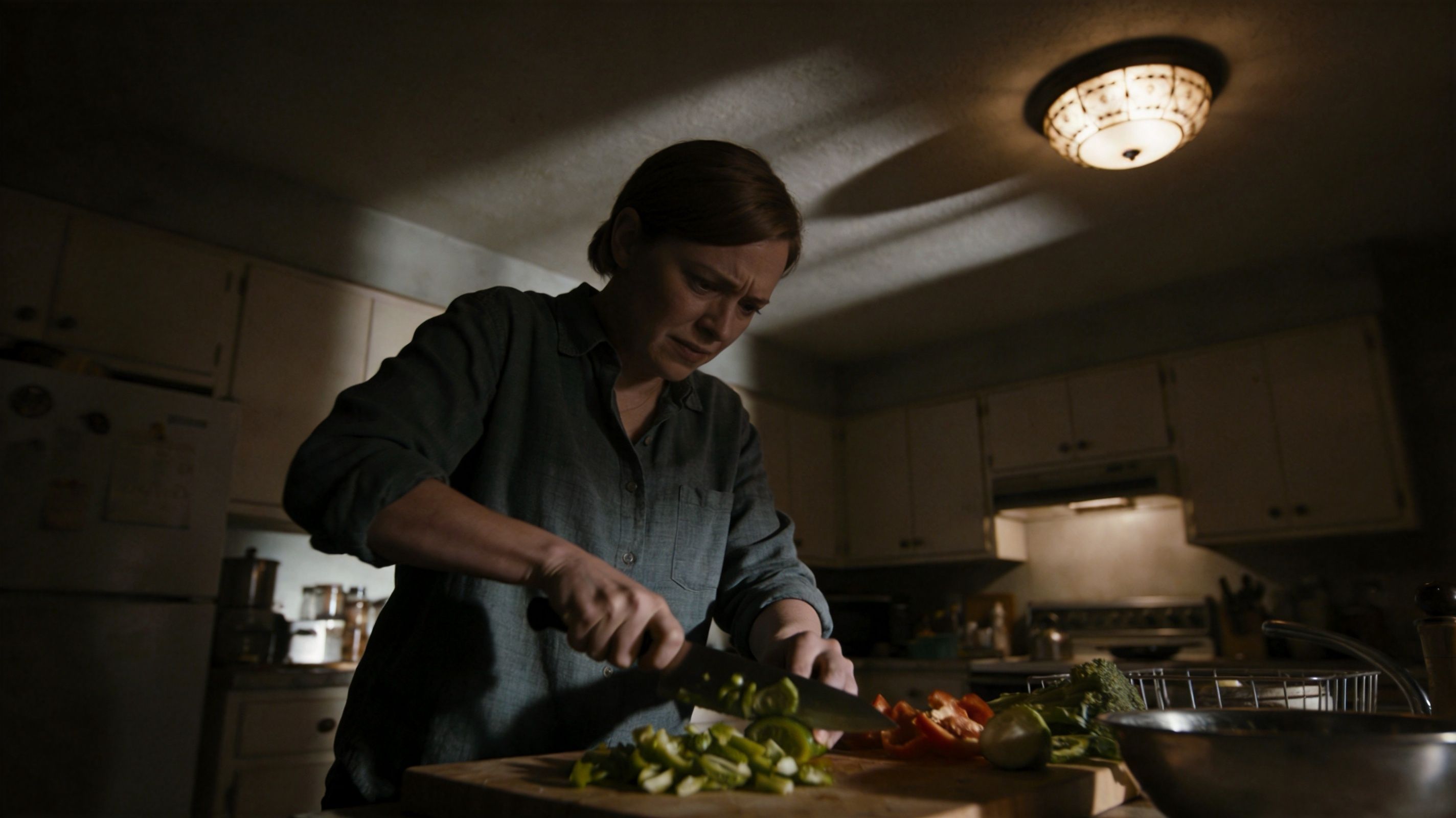 A Woman With A Concerned Expression Prepares Vegetables In A Dimly Lit Kitchen With Overhead Lighting.