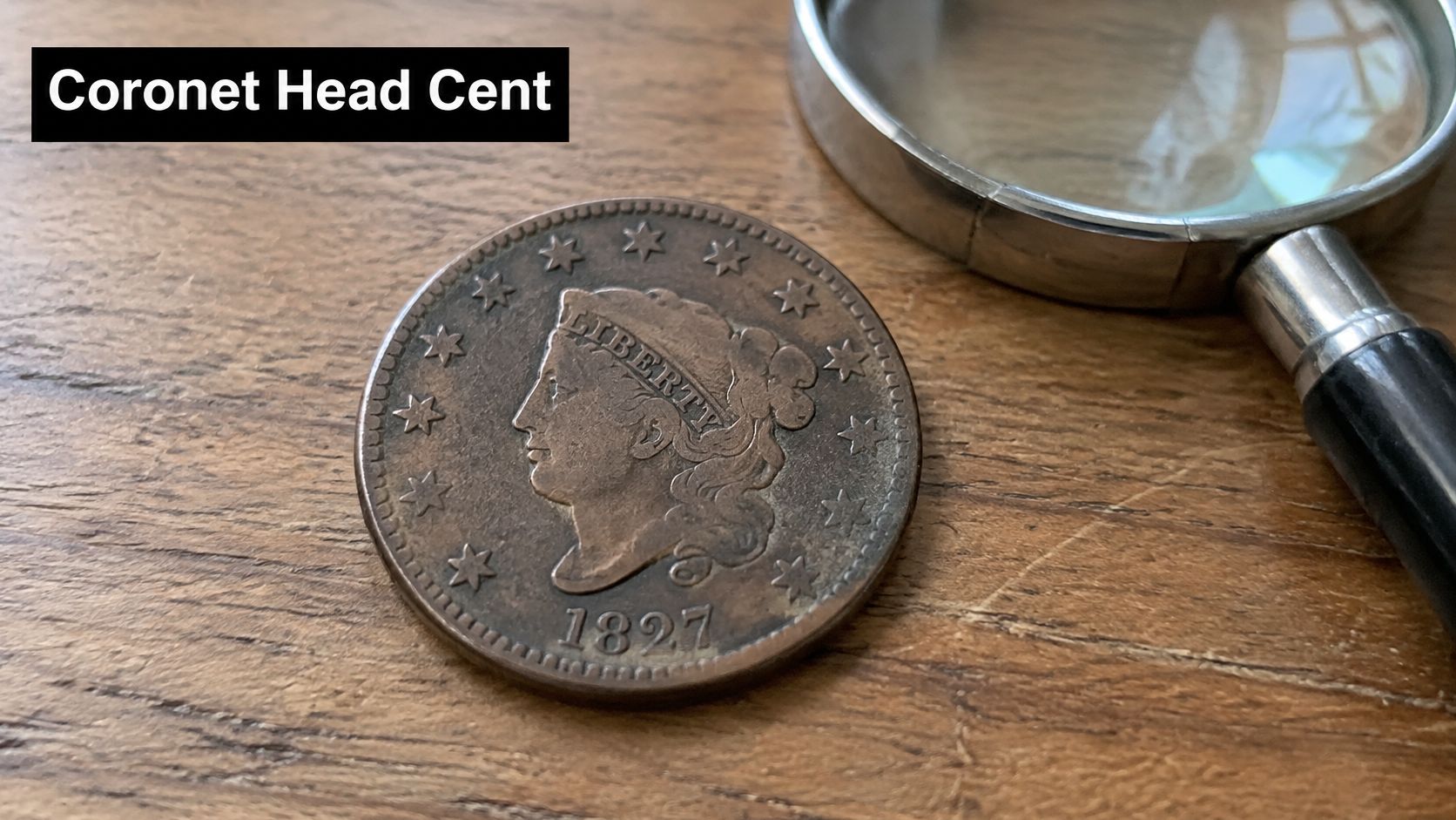 An 1827 Coronet Head Large Cent coin sitting on a wooden surface next to a magnifying glass.