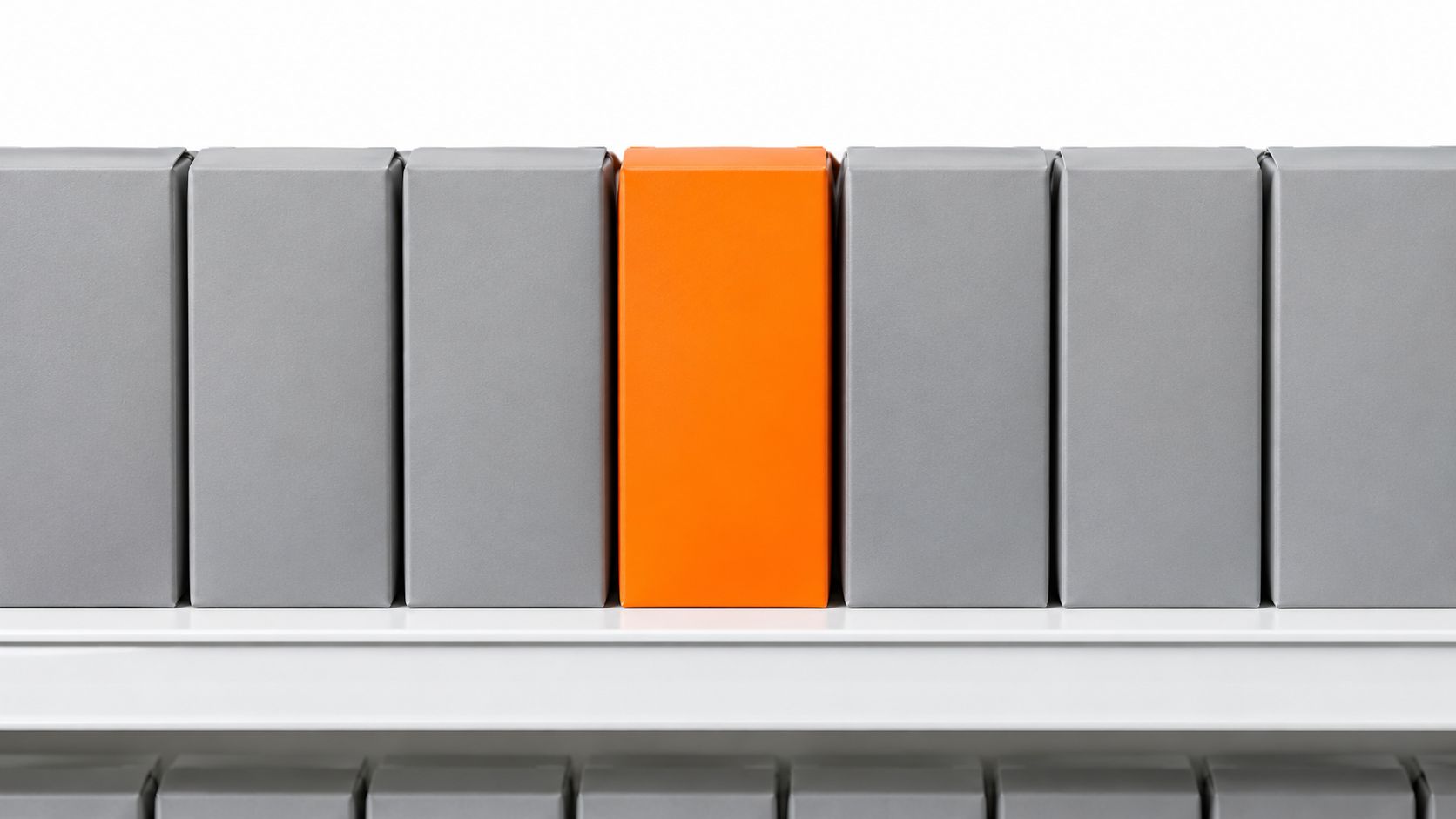 Eine einzelne leuchtend orangefarbene Verpackung steht hervorstechend zwischen mehreren schlichten grauen Boxen in einem Verkaufsregal.