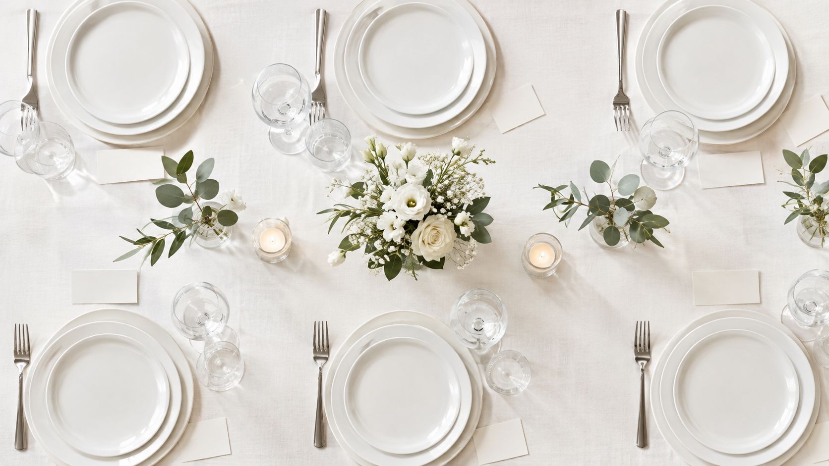 Mesa elegante preparada para un banquete con vajilla blanca, flores frescas, velas y cubiertos plateados sobre mantel.