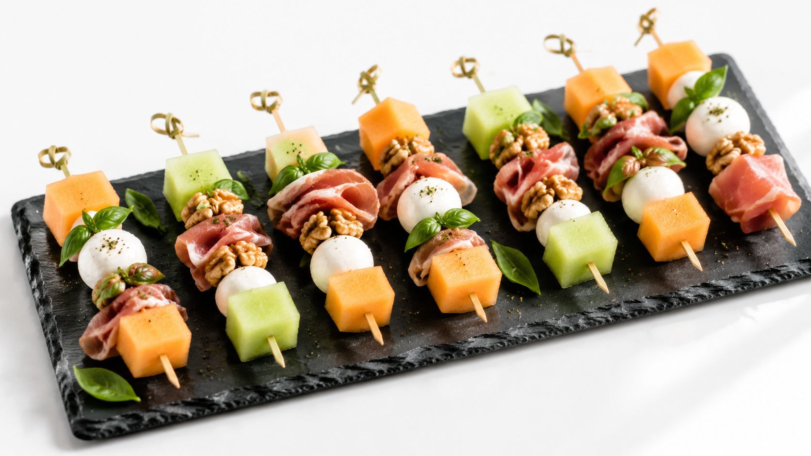 Brochetas gourmet de melón con jamón serrano, queso mozzarella y nueces sobre una bandeja de piedra negra.