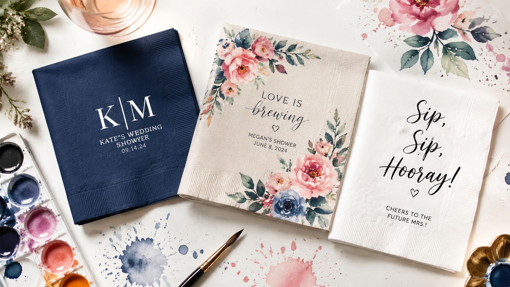 Three custom wedding shower napkins with floral and monogram designs displayed next to watercolor paint and brushes.