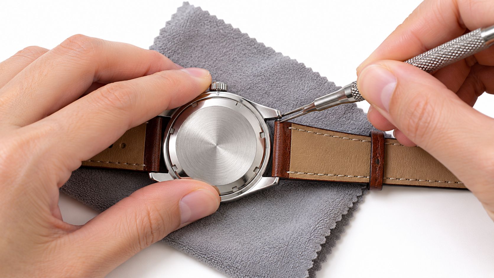 A pair of hands using a spring bar tool to remove a brown leather watch strap.