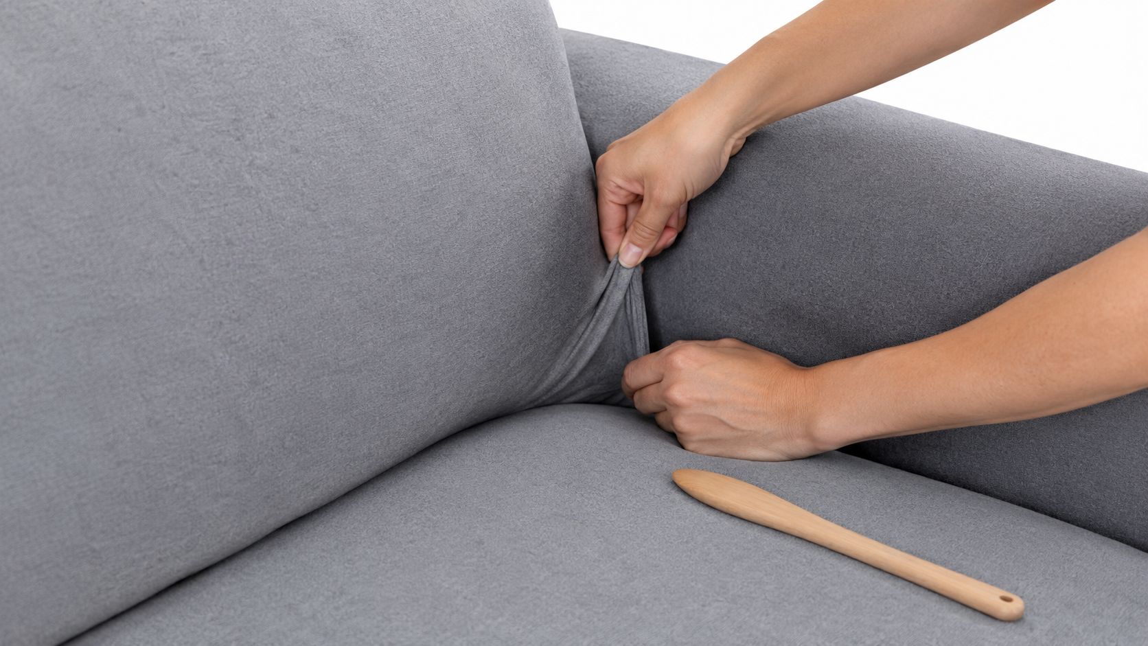 A person tucking a grey stretch sofa cover into the crease beside a sofa arm, with a wooden tool on the seat.