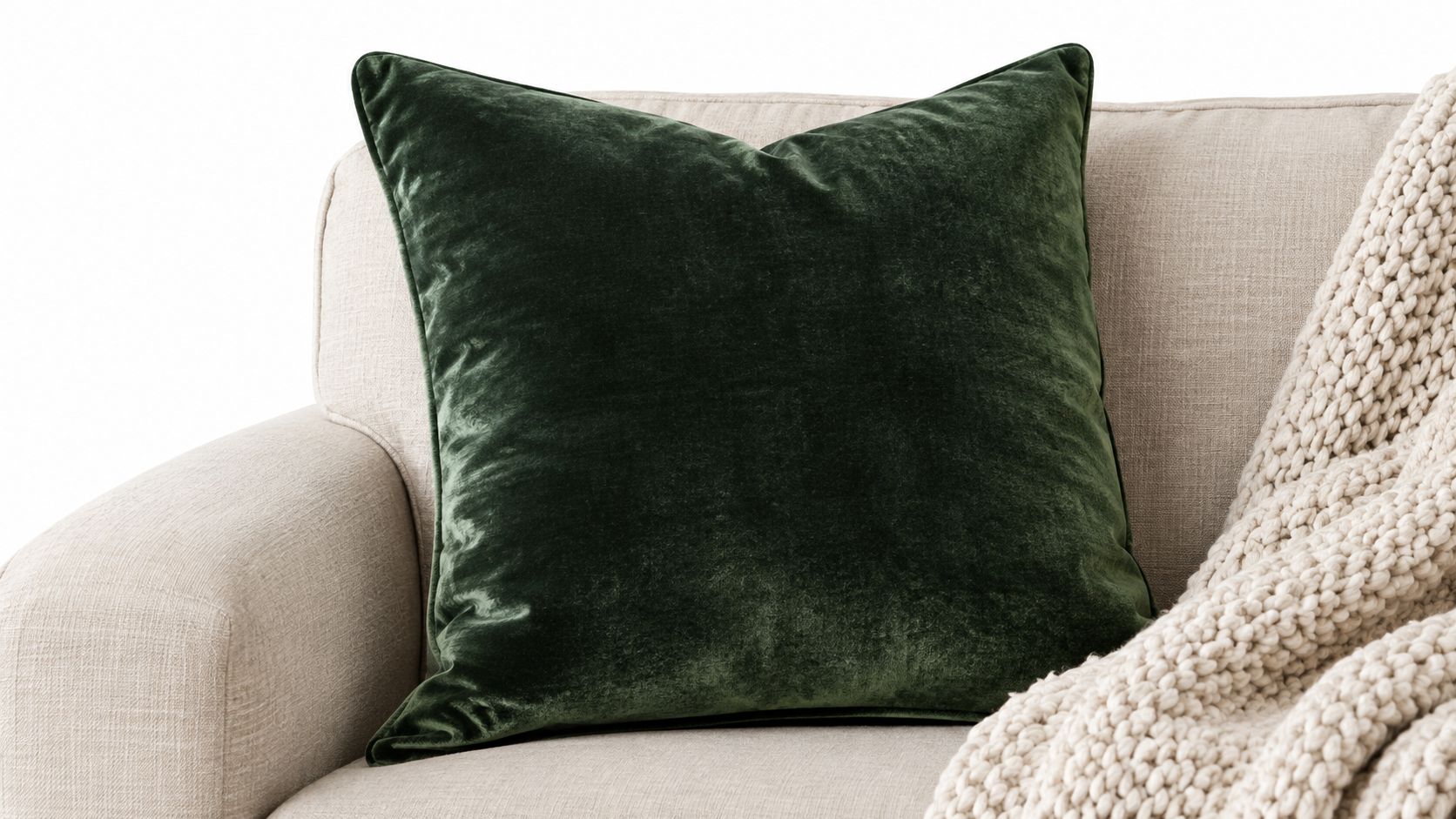 A plush dark green velvet decorative square cushion resting on a beige sofa with a knit blanket.
