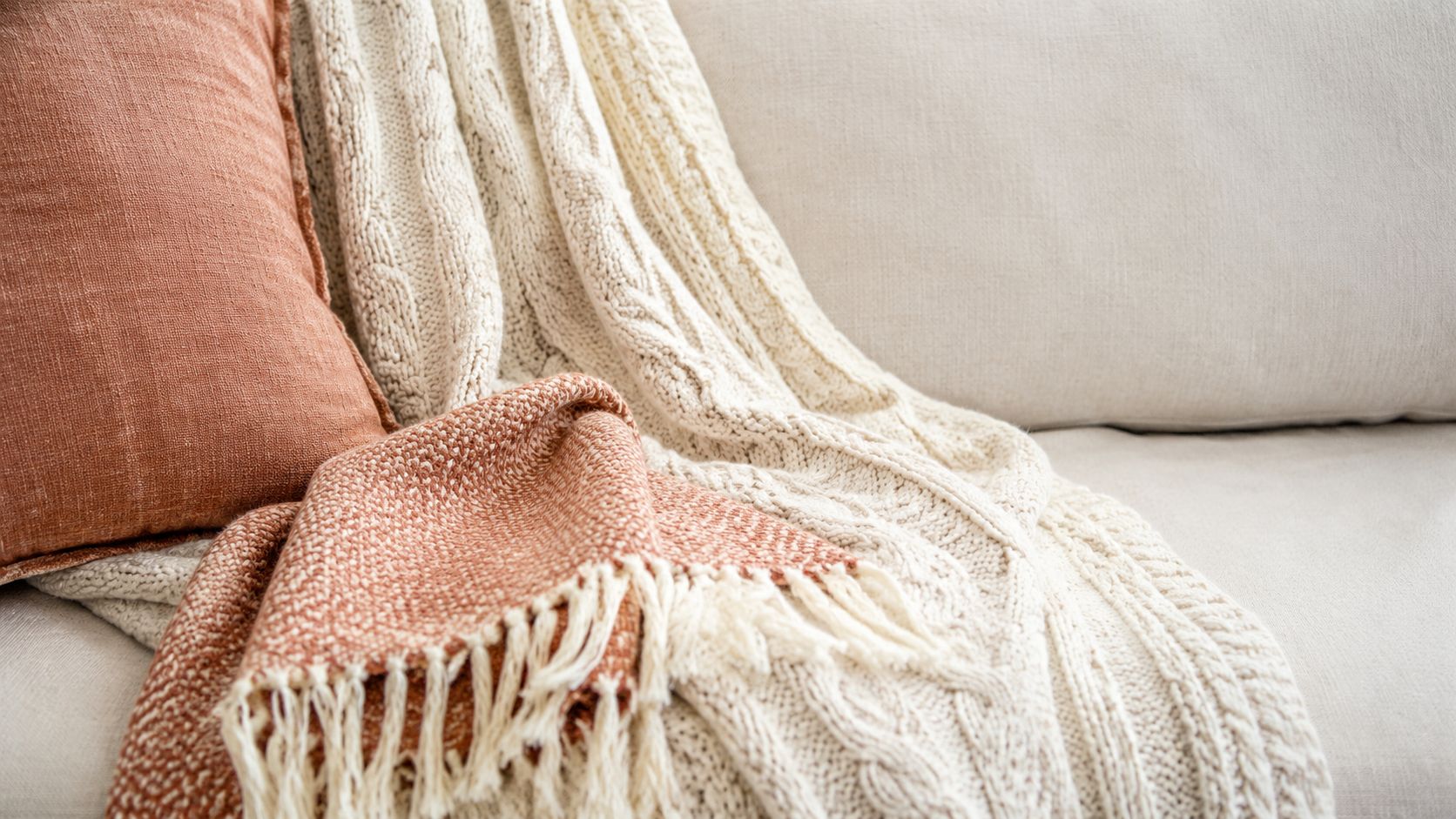 A cozy beige couch features a textured cream knit blanket and a warm orange throw pillow