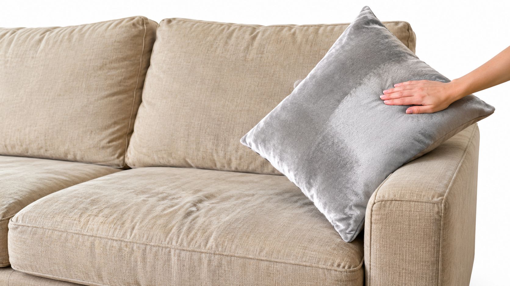 A hand places a soft grey velvet cushion onto a neutral beige fabric sofa.
