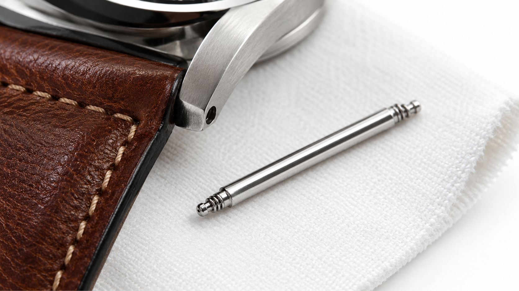 A steel spring bar rests on a white cloth next to a leather watch strap and case.