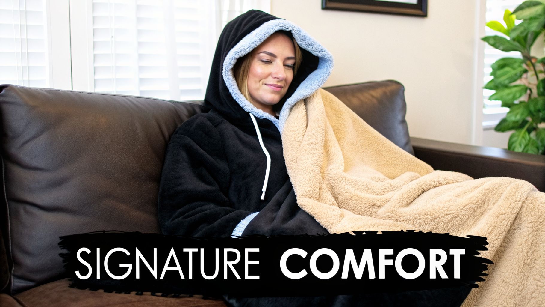 Woman relaxing on a brown couch wearing a black oversized hooded blanket with a beige throw.