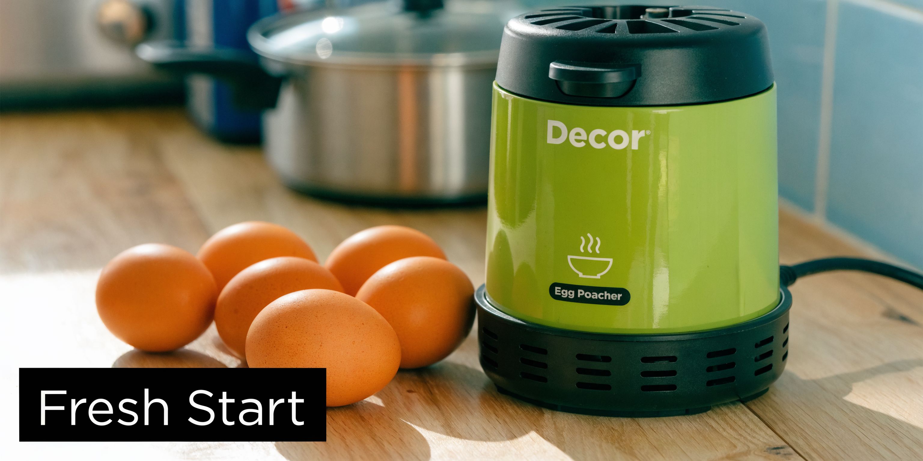 A green Decor electric egg poacher sits on a wooden counter next to several fresh brown eggs.