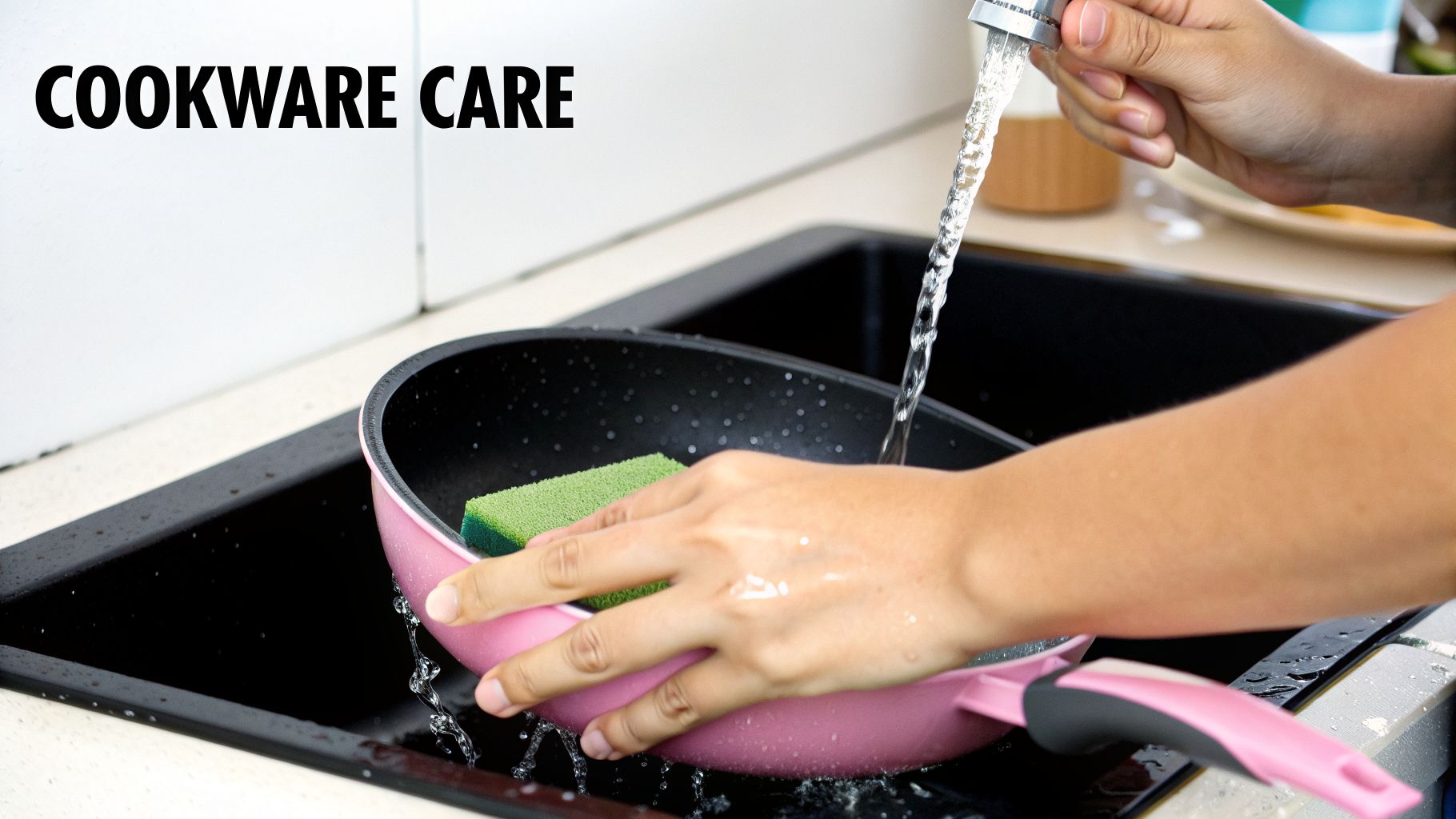 A person's hands washing a pink frying pan with a green sponge under running water in a black kitchen sink.