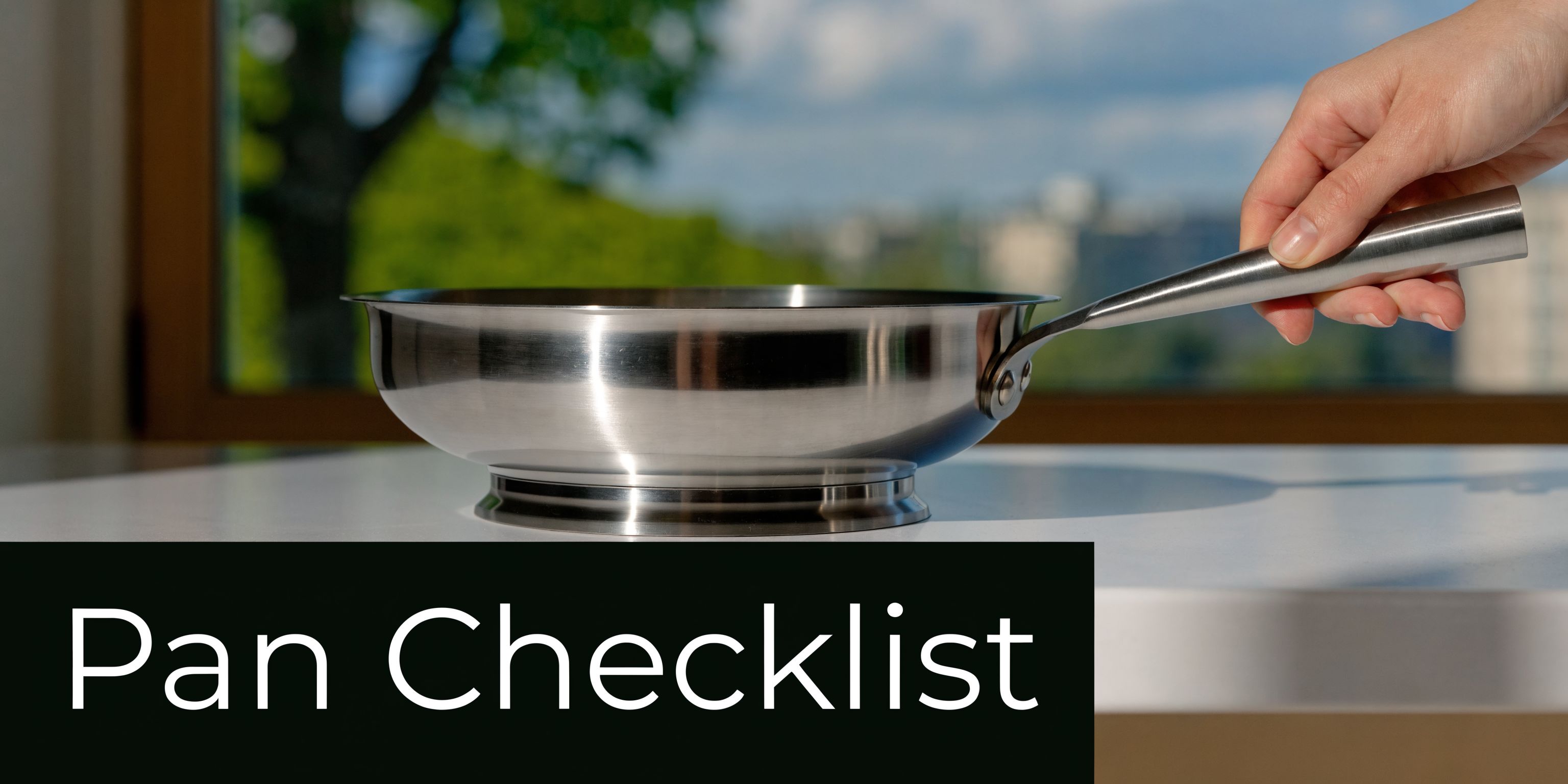 A hand holds a stainless steel saute pan on a white table with a blurred background
