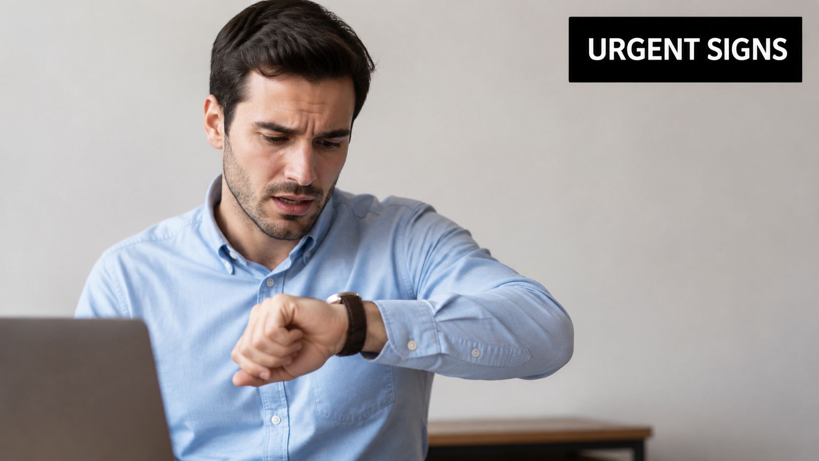 A concerned man in a blue shirt checks his watch while working on a laptop computer