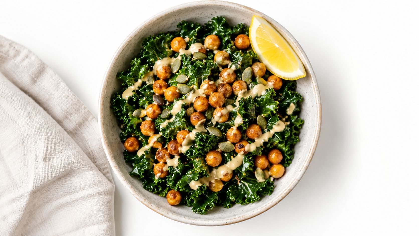A healthy kale salad in a ceramic bowl topped with crispy roasted chickpeas, pumpkin seeds, and tahini dressing.