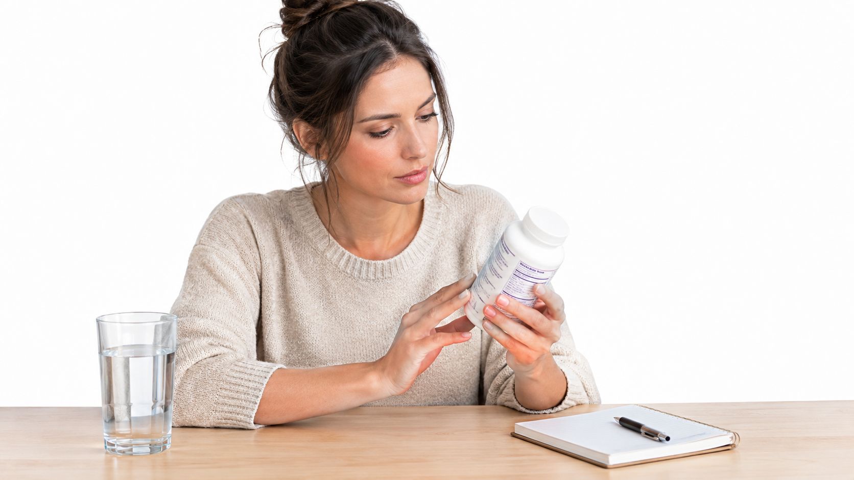 A woman holding a bottle of prenatal vitamins while sitting at a wooden table with water.