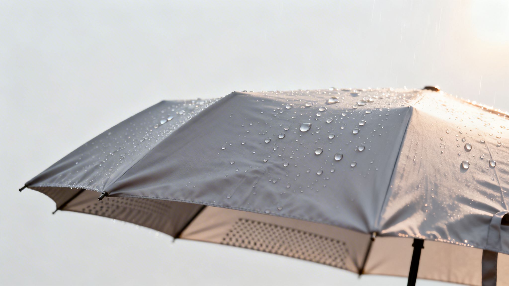 Close-up of a grey umbrella covered in glistening raindrops against a bright, hazy sky, suggesting light rain.
