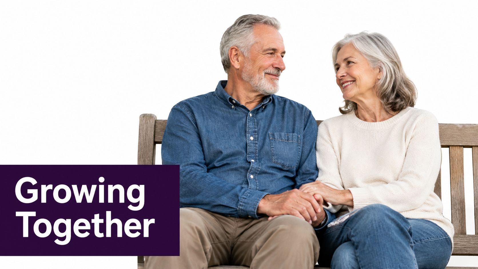 A happy senior couple sitting on a wooden bench, smiling and holding hands, looking at each other.