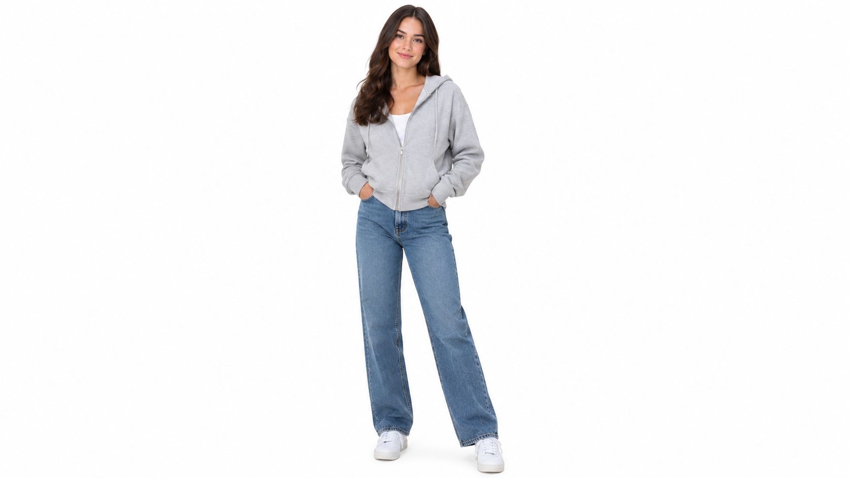 A young woman wearing a light grey zip up hoodie, jeans, and white sneakers stands smiling.