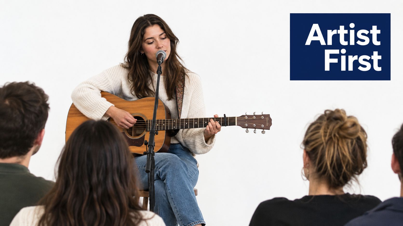 A female musician plays an acoustic guitar and sings in front of a small seated audience.