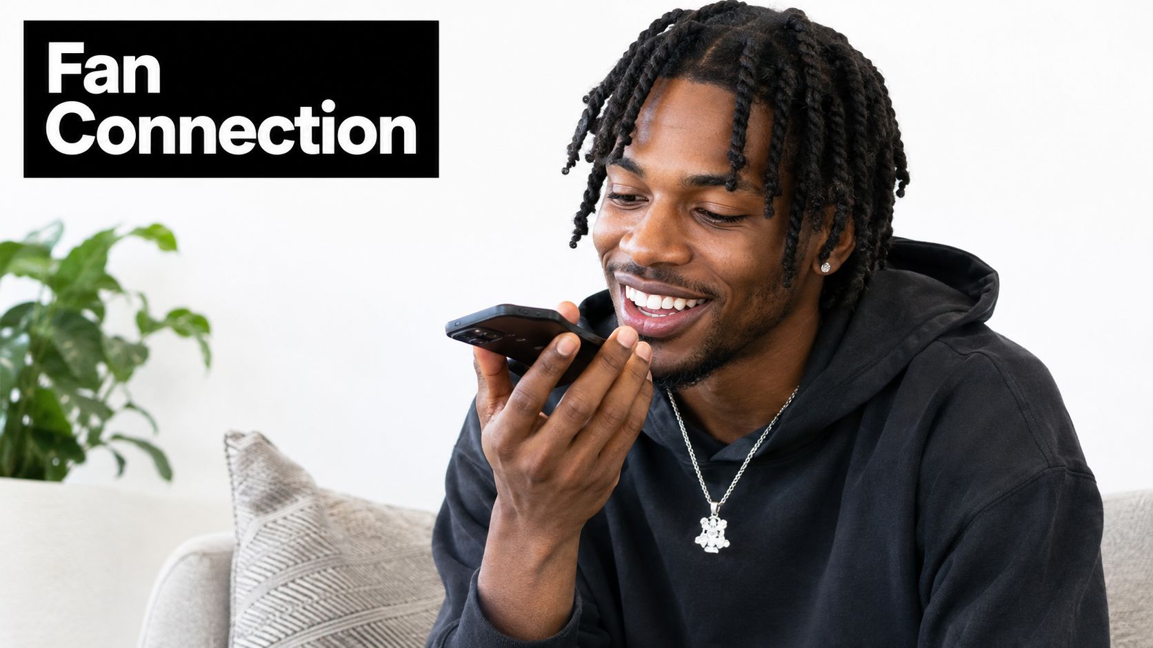 A smiling young man with braided hair talking on a smartphone while sitting on a couch.