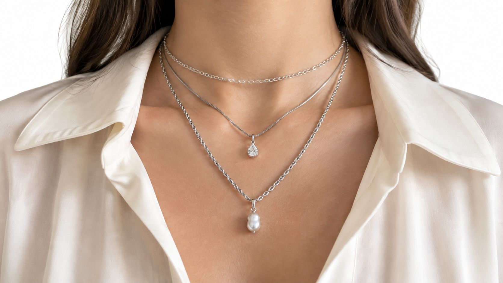 A woman wearing three layered silver necklaces, featuring a teardrop pendant and a pearl charm.
