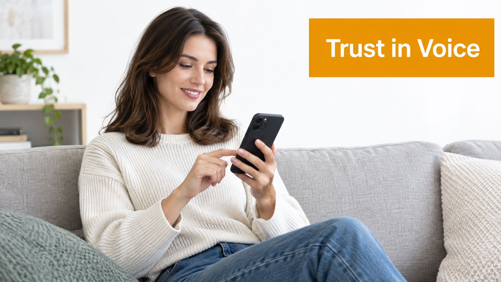 A smiling young woman sitting on a couch while using a smartphone in a bright living room.