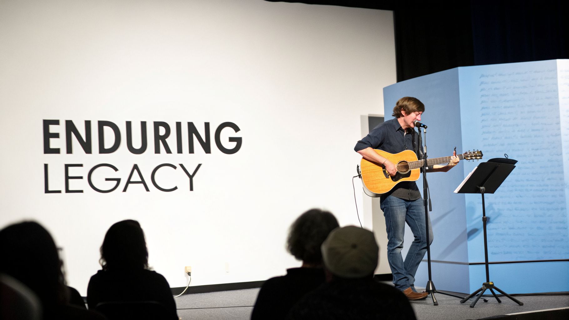 A man plays acoustic guitar and sings on stage with "ENDURING LEGACY" projected behind him.