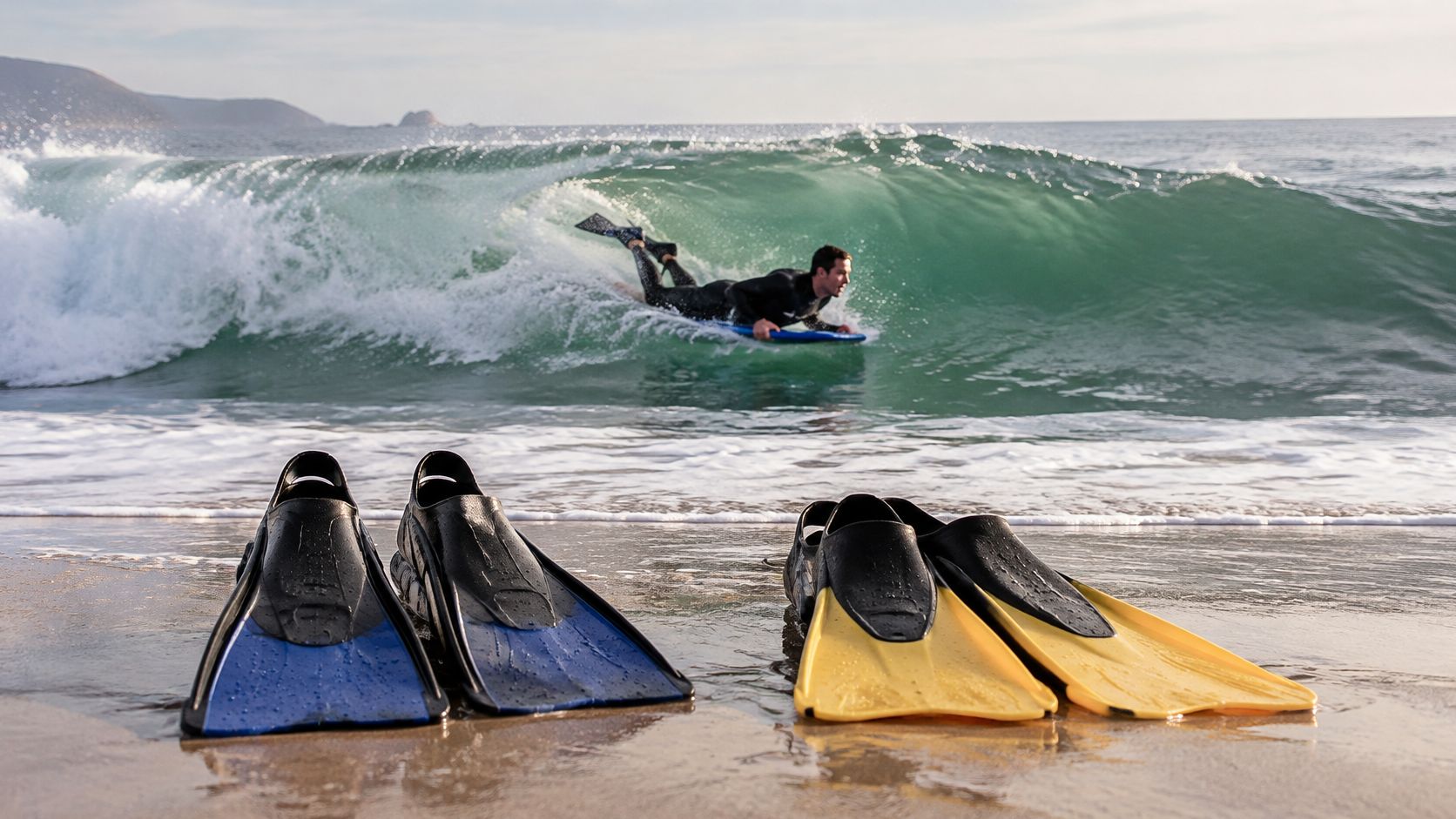 A man bodyboarding on a wave behind two pairs of swim fins placed on the sandy beach.