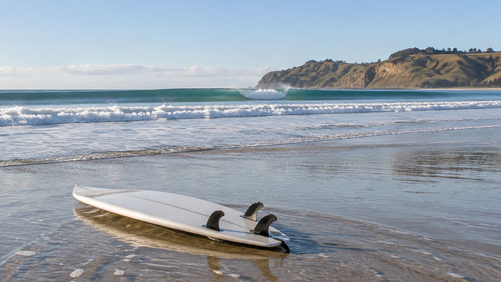 A white surfboard with black fins rests on the wet sand of a scenic beach at low tide.