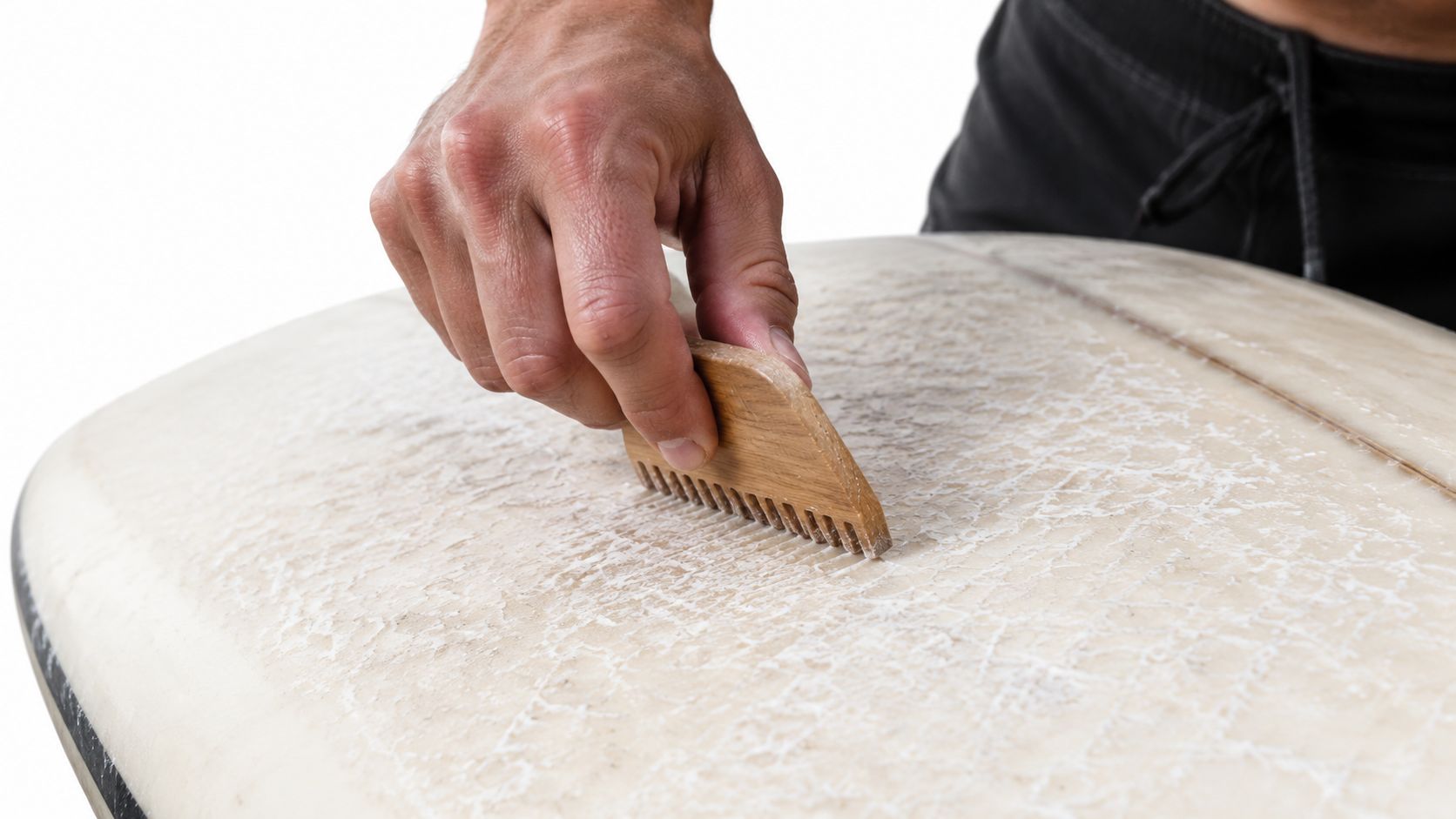 A surfer using a wooden wax comb to remove old surf wax from a surfboard surface.