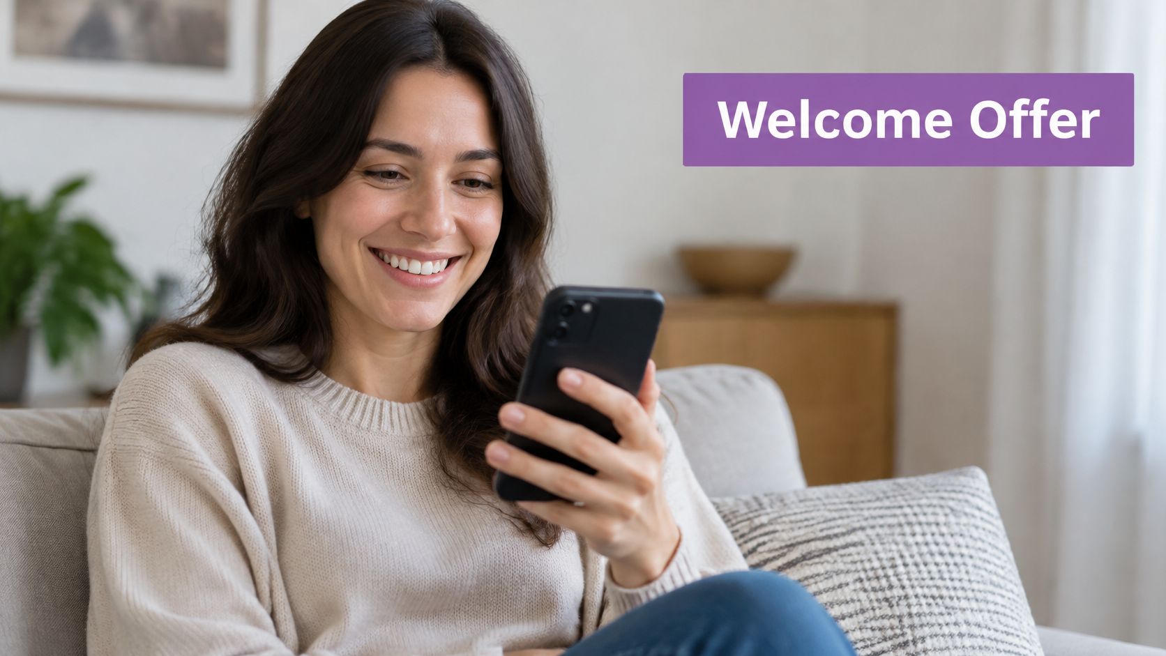 A smiling young woman sitting on a sofa while reading a welcome offer on her smartphone.