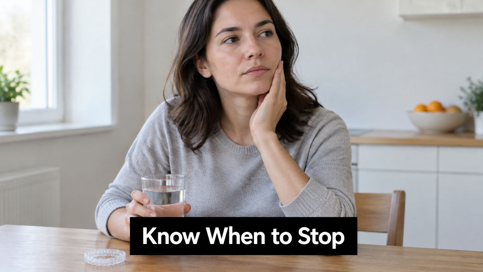 A woman holding a glass of water while touching her cheek, sitting at a wooden table.