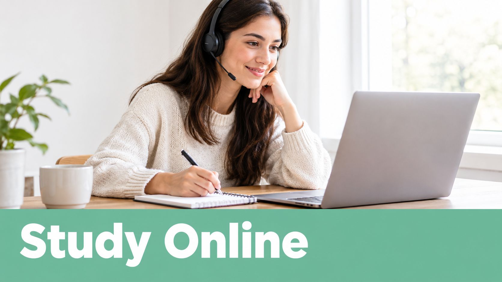 A smiling young woman wearing headphones and taking notes while studying online on her laptop at home.