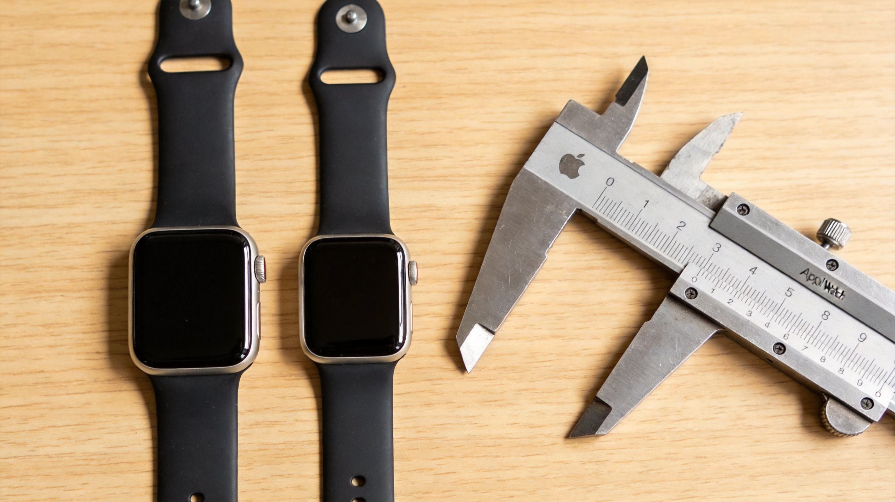 Δύο ρολόγια Apple Watch δίπλα σε ένα μεταλλικό παχύμετρο πάνω σε μια ξύλινη επιφάνεια.