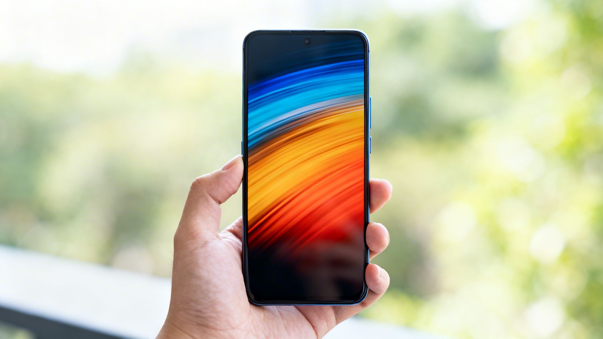 Χέρι κρατά ένα μπλε smartphone με πολύχρωμη ταπετσαρία μπροστά σε θολό πράσινο φόντο.