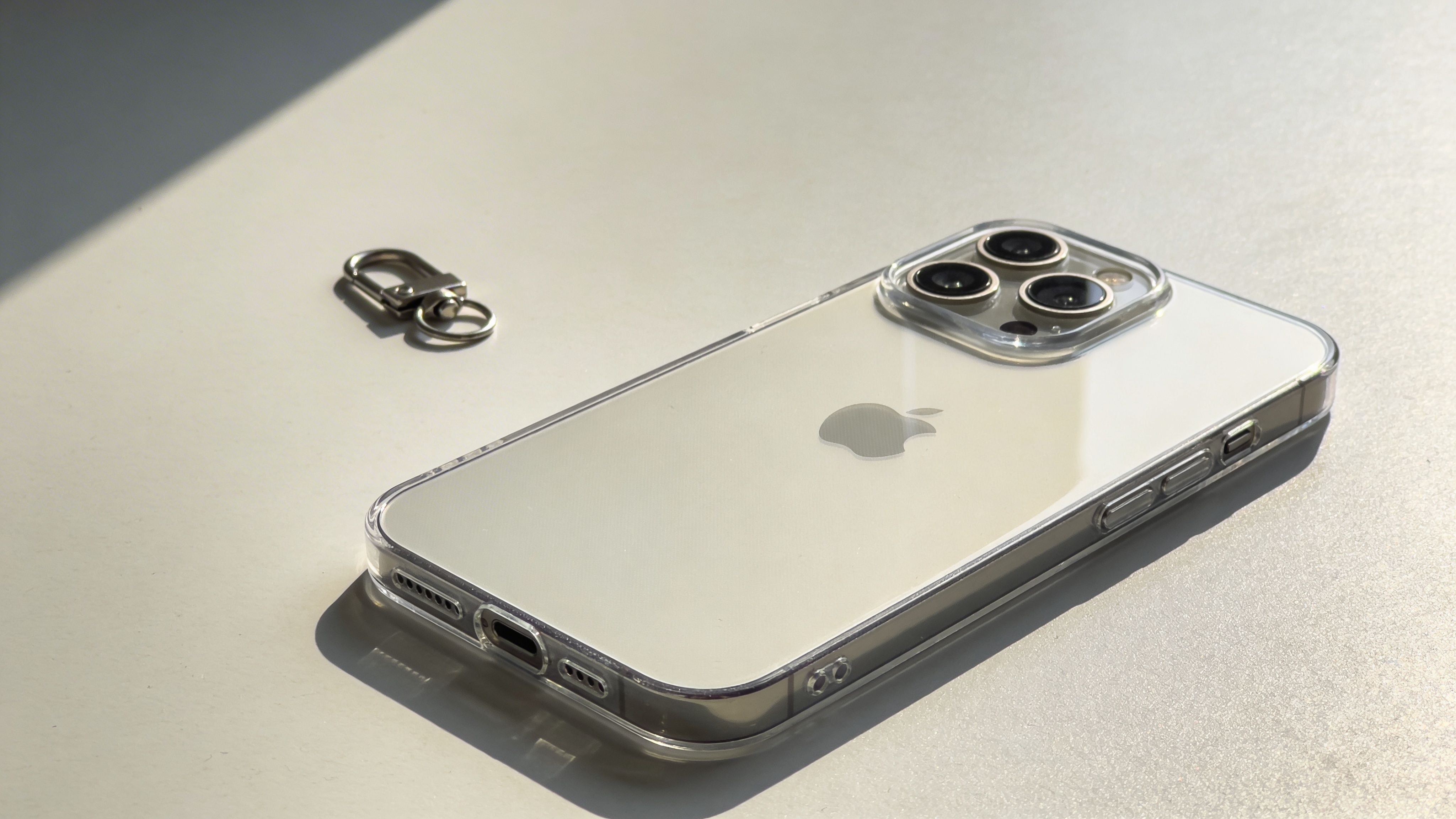 Ένα λευκό iPhone 14 Pro με διάφανη θήκη προστασίας τοποθετημένο δίπλα σε ένα μεταλλικό κρικάκι κλειδιών σε λευκό φόντο.