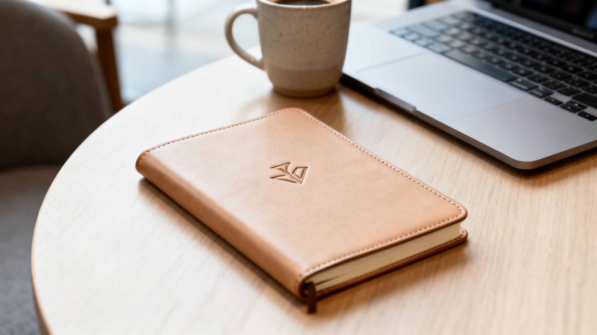 A light brown leather-bound notebook with an embossed logo rests on a wooden table with a coffee and laptop.