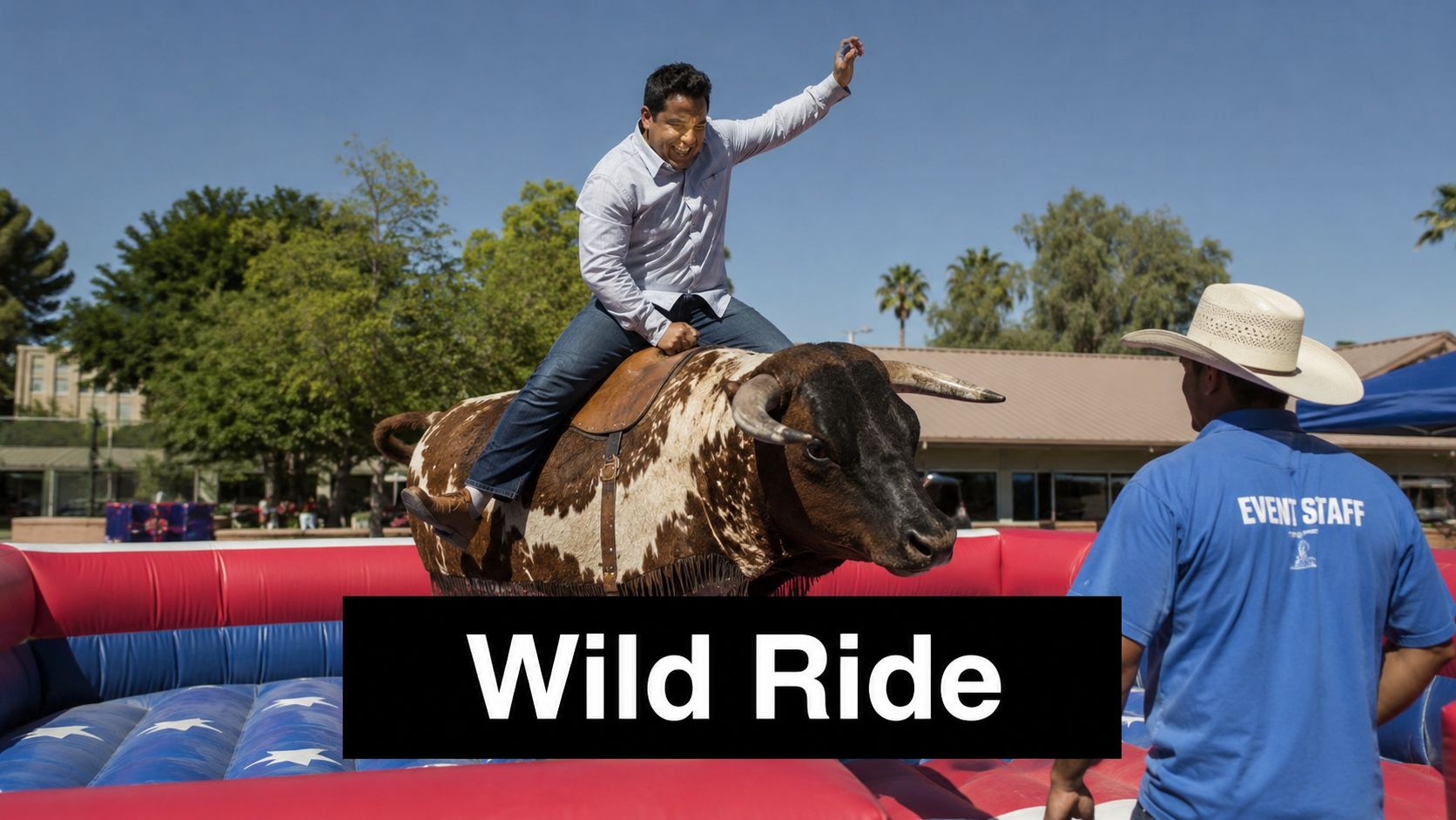 A man enthusiastically riding a mechanical bull at an outdoor event while event staff stands nearby.
