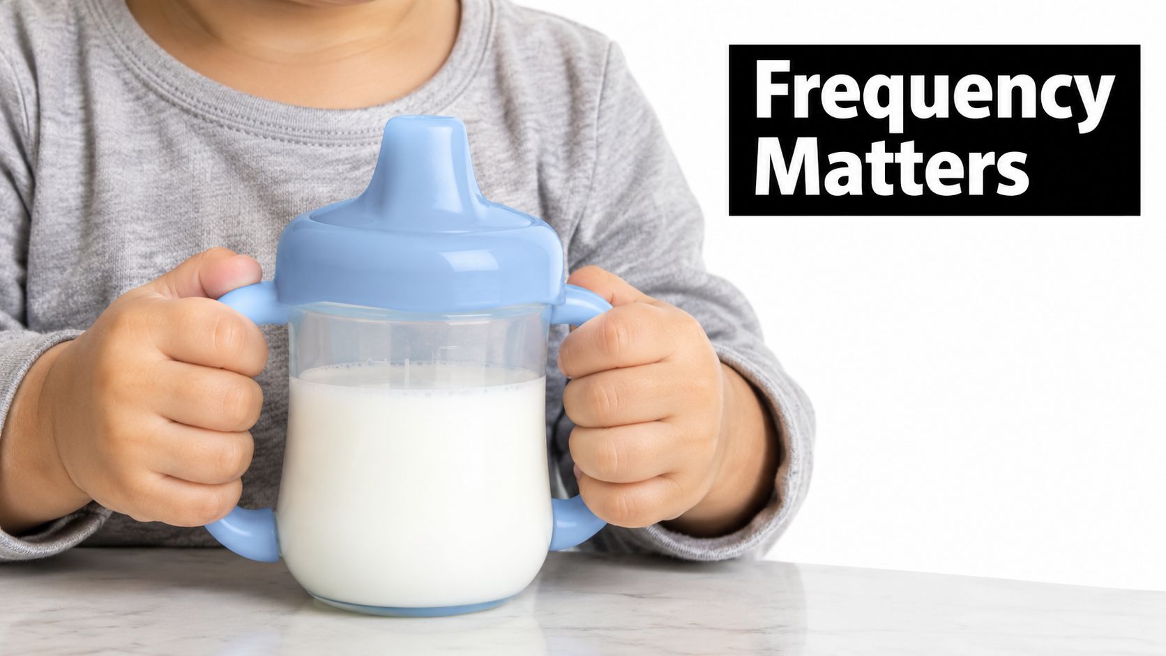 A toddler holding a sippy cup filled with milk while sitting at a table.