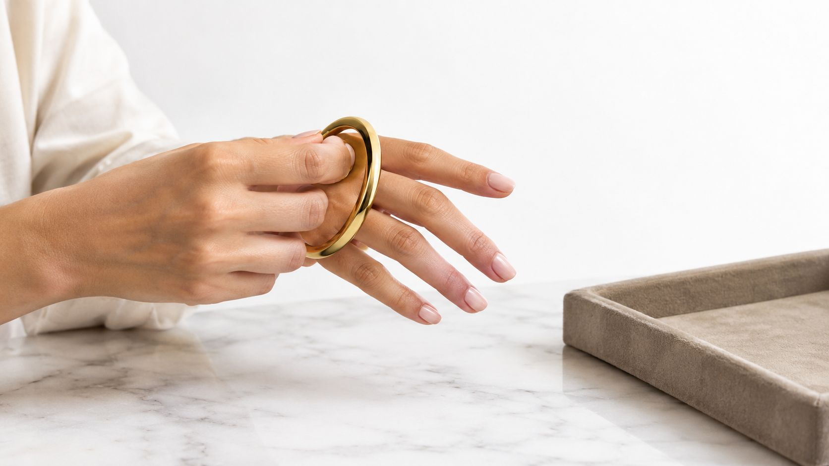 A person demonstrating how to wear a smooth gold bangle bracelet on their wrist at a table.