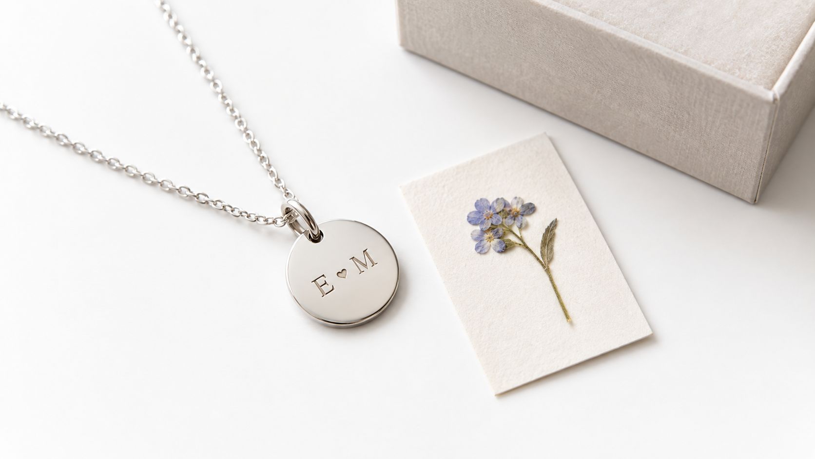A silver engraved necklace with a heart symbol and E M letters next to a pressed flower.